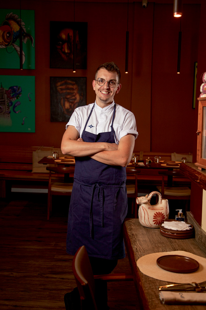 O chef Bruno Hoffman posa de avental azul marinho no ambiente do restaurante o qual comanda, o Caos Brasilis