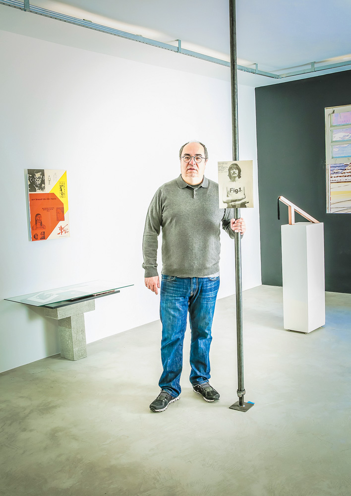 Um homem grisalho de meia idade posa em uma galeria de arte. Ele segura um cabo que tem uma foto antiga pendurada