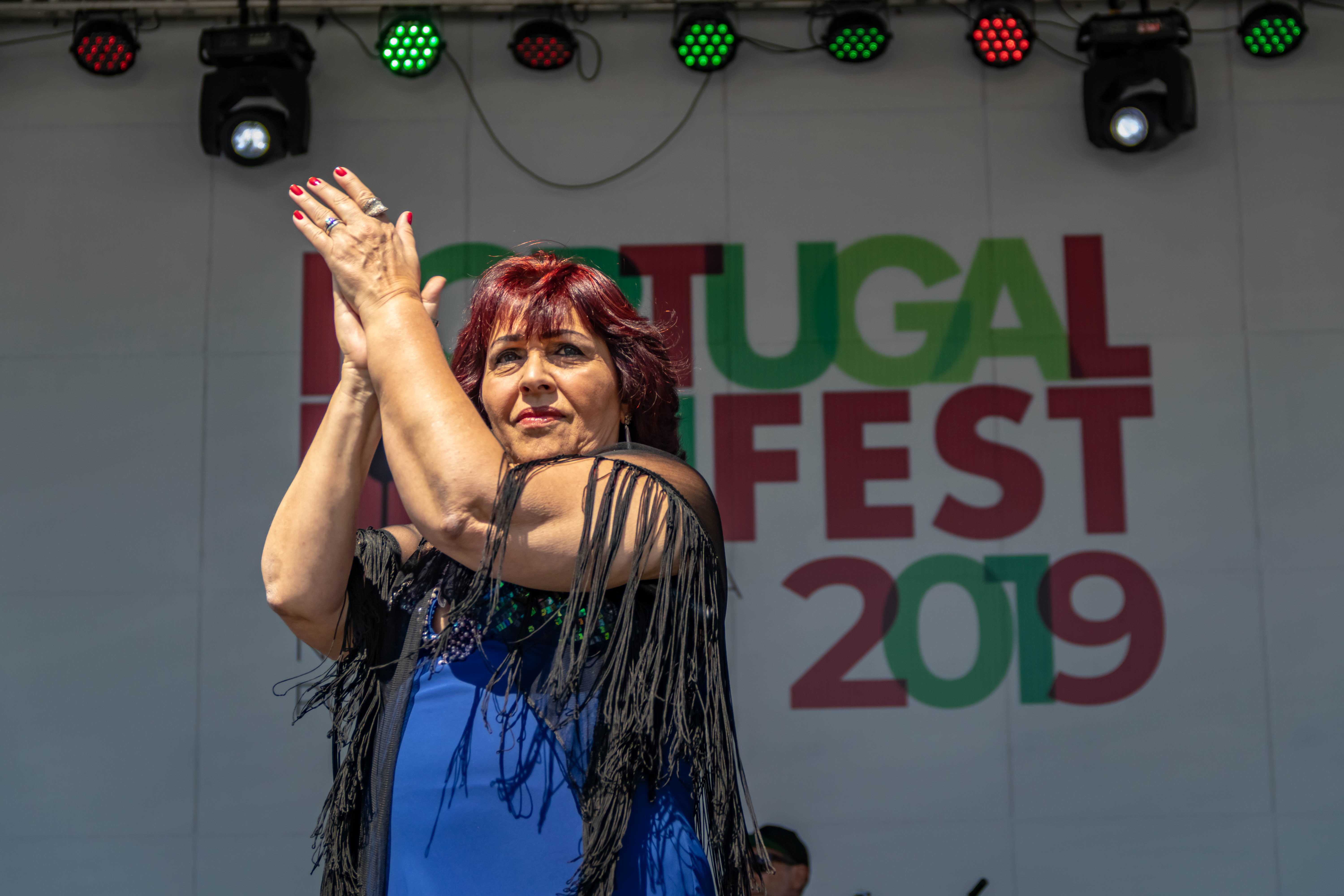 Ciça Marinho se apresenta no palco do Portugal Fest 2019. Ela é uma mulher branca, de cabelos vermelhos e veste uma roupa azul com franjas pretas.