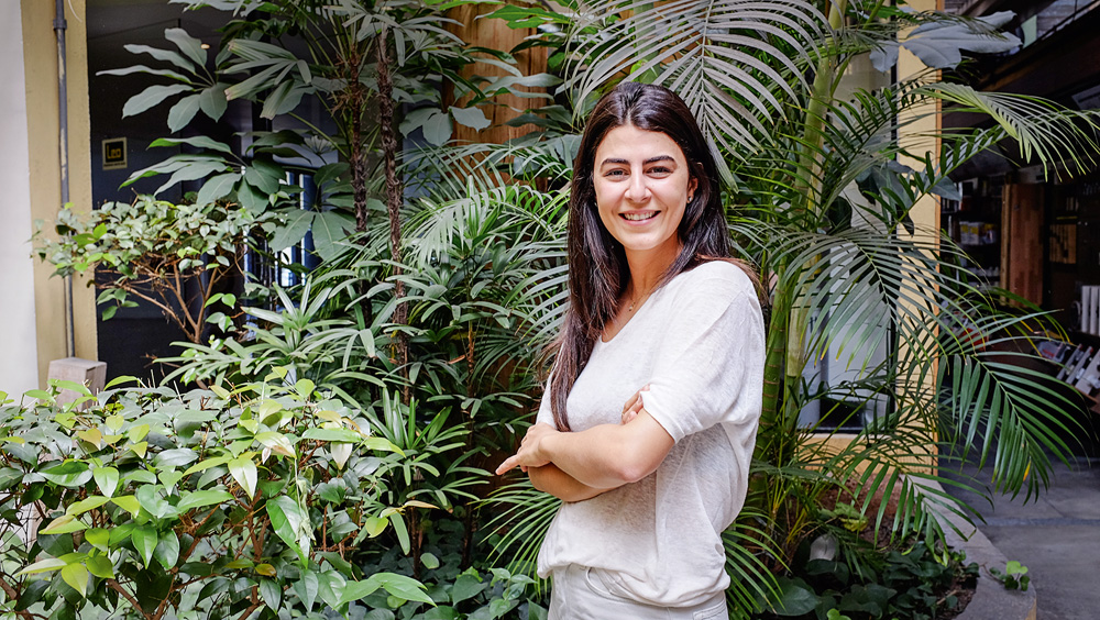 A imagem mostra Marcella, de braços cruzados, em frente a um lugar cheio de plantas verdes.