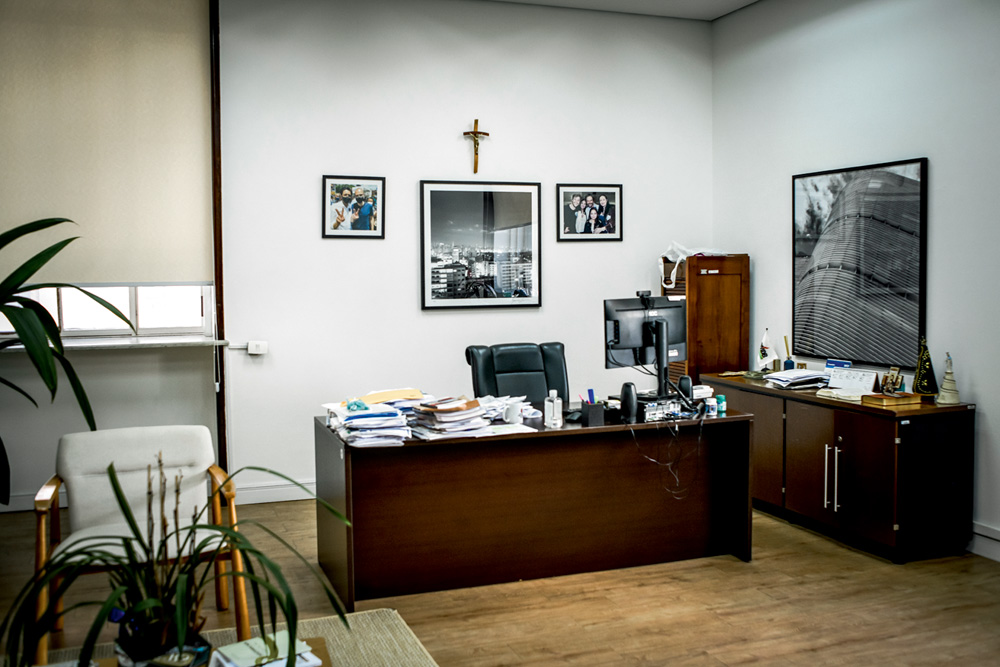 Sala de Ricardo Nunes. Tem uma mesa com várias papeladas, quadros pequenos na parede, uma cruz, e armários