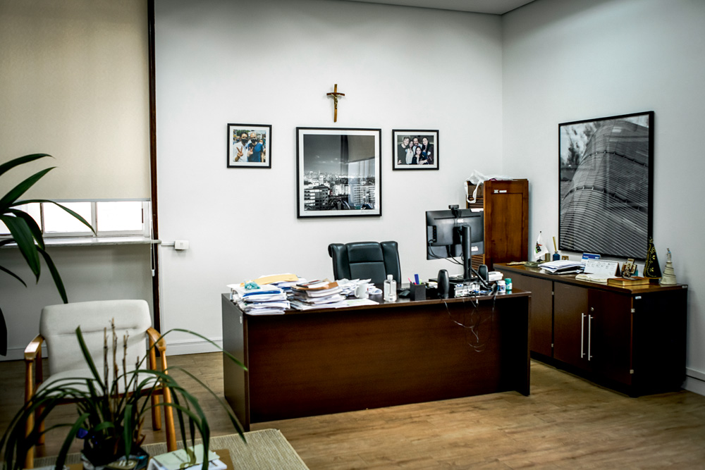 RICARDONUNES_21.JPG Sala de Ricardo Nunes. Tem uma mesa com várias papeladas, quadros pequenos na parede, uma cruz, e armários