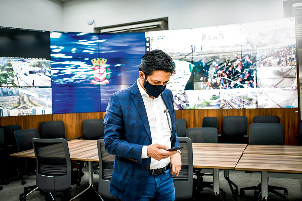 O prefeito Ricardo Nunes está em uma sala com várias televisões que transmitem imagens da cidade. Ele está mexendo no celular