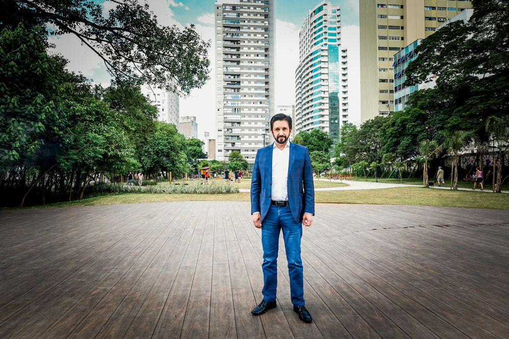 O prefeito Ricardo Nunes posa de pé no Parque Augusta, ao fundo, se vê prédios. Ele veste terno azul e calça jeans