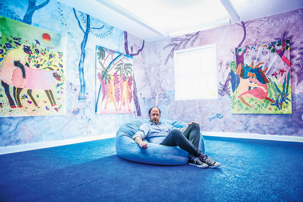 Em uma sala de exposição em tons de azul e roxo, há um homem deitado em um puff. Nas paredes, artes de árvores e plantas. Há também quadros coloridos que parecem remeter a animais e natureza