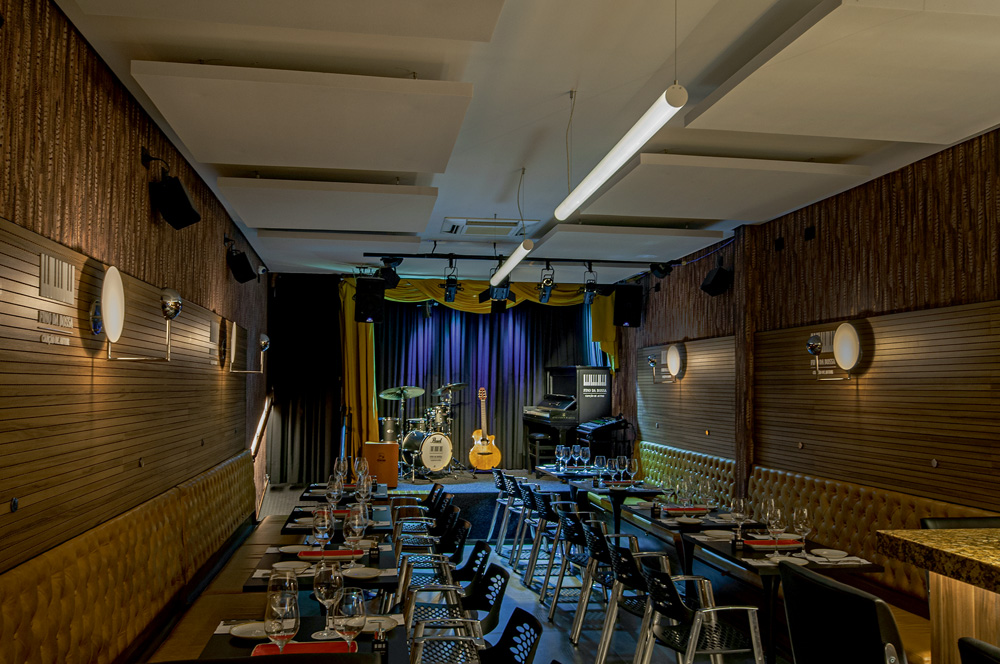 Imagem mostra espaço de restaurante e palco ao fundo com um piano, um violão e uma bateria. Várias mesas e cadeiras preenchem o espaço até o palco, que conta com uma luz azulada.