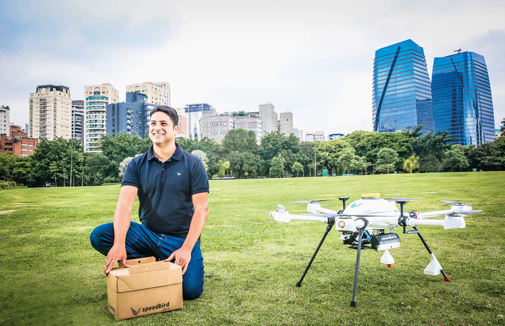 A imagem mostra Salomão, segurando uma caixa, e do lado dele, um drone estacionado na grama. Ele está em um ambiente aberto.