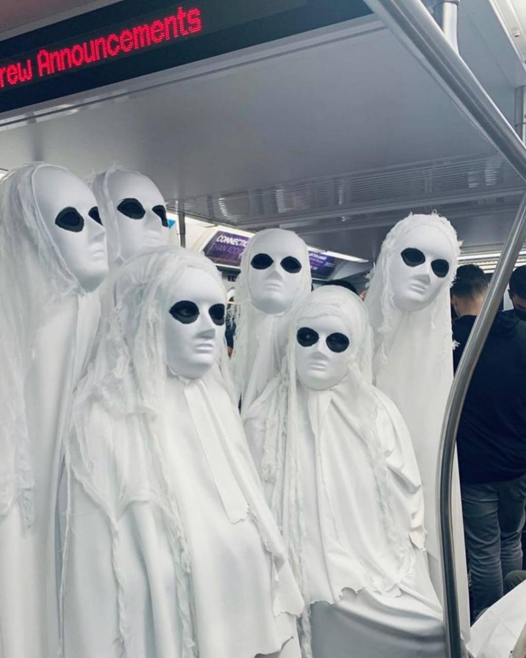 A imagem mostra seis pessoas em fantasias de fantasma. Estão vestidas inteiras com um pano branco e nos olhos há uma maquiagem preta.