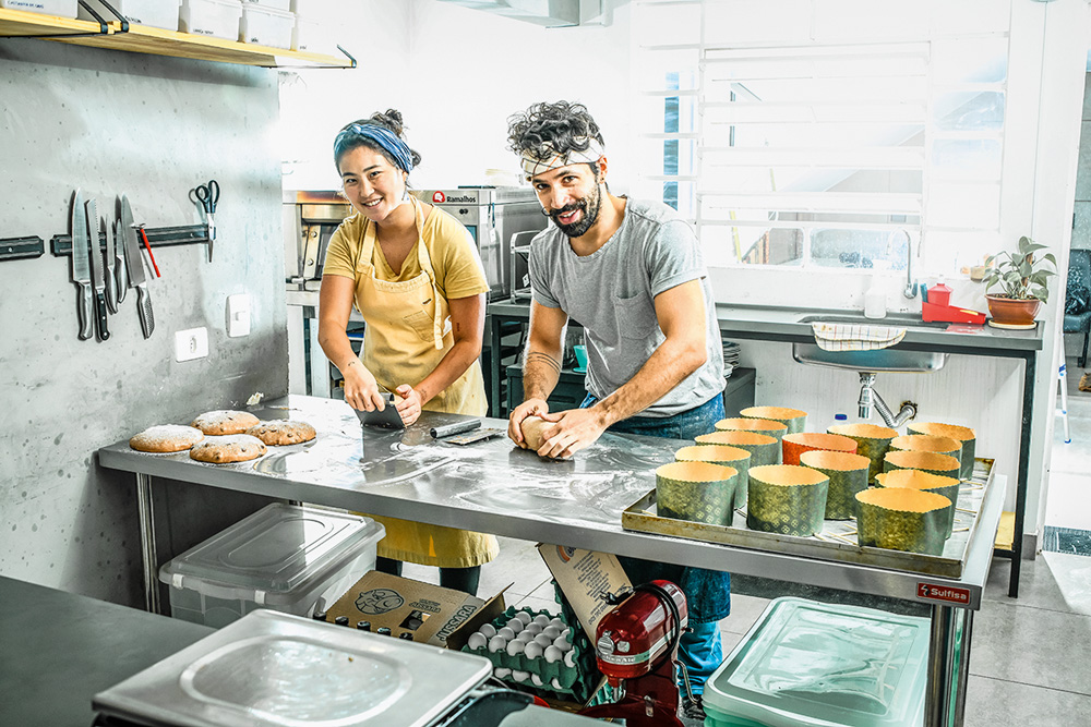 Um homem e uma mulher estão em uma cozinha, sorrindo para a câmera, fazendo massas. Tem cookies prontos na tábua e formas com alimentos