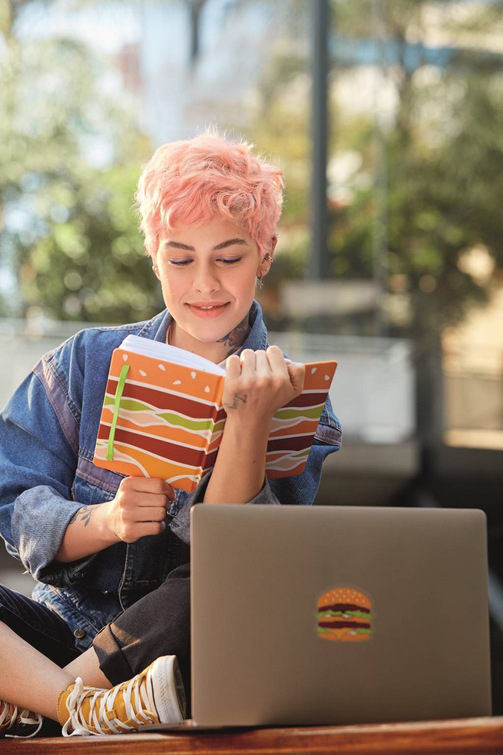 A imagem mostra uma mulher com um caderno com estampa de hamburguer, além de um notebook que também possuí uma estampa de hambúrguer.