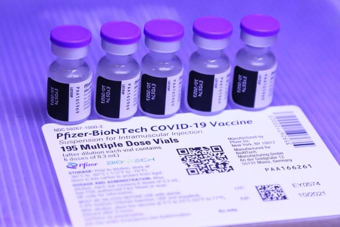 Imagem mostra 5 frascos de vacina Pfizer colocados sobre a bula do composto. O fundo é de cor azul e roxa.