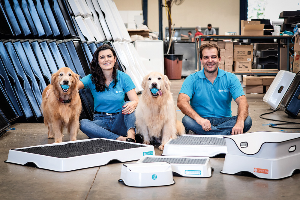 A imagem mostra Lara e Fabio, ambos sentados no chão, ao lado de dois cachorros. Na frente deles, os tapetes e outros itens que criaram para os pets.