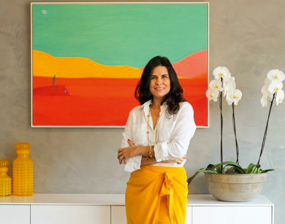 Imagem mostra muler de camisa branca e saia amarela posando com os braços cruzados em frente a uma parede cinza com um quadro colorido e um vaso de orquídea.