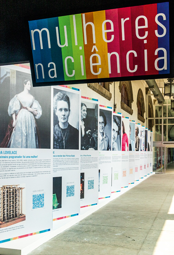 Em um corredor, estão enfileirados grandes banners com fotos e informações sobre diferentes mulheres cientistas. Há, no início do corredor, uma placa escrito "Mulheres na Ciência"