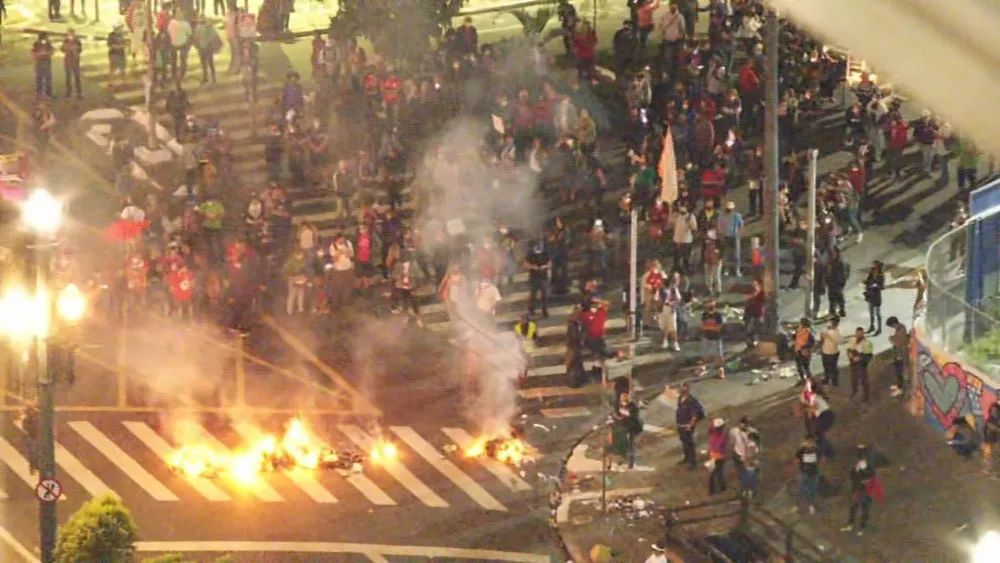 Uma foto aérea mostra um grupo de manifestantes em um cruzamento. Há sacos de lixo pegando fogo e fumaça subindo.