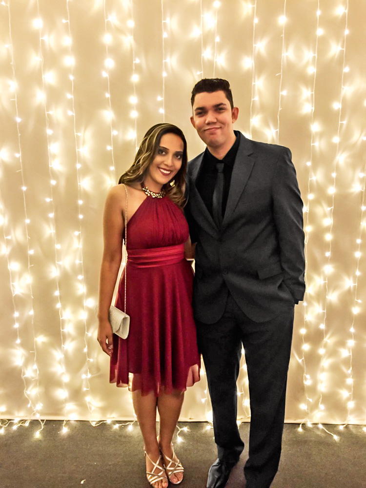 Um casal, a mulher de vestido vermelho e o homem de terno preto, posam em frente a uma parede branca toda iluminada com pequenas luzes.