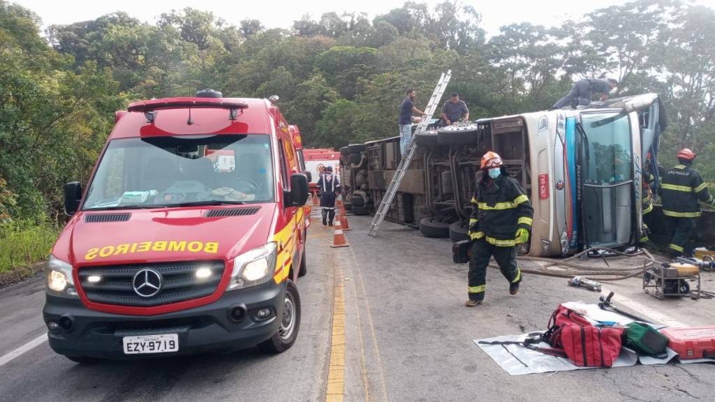 Imagem mostra ônibus tombado em lateral de pista e, ao lado, viatura dos Bombeiros. Bombeiros circulam pelo local e uma escada foi colocada para subir no ônibus