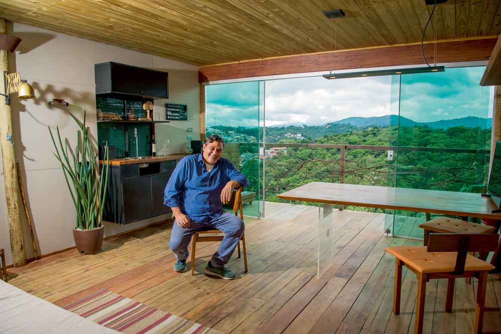 A imagem mostra Ricardo Afiune sentado em um banco de madeira em uma sala repleta de móveis de madeira. Ao fundo, uma varanda que dá vista em um lugar cheio de árvores e verde.