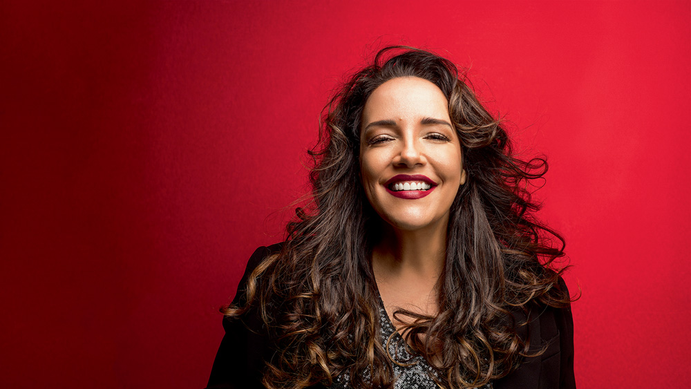 ana carolina sorrindo para a câmera com cabelo solto, batom vermelho escuro. o fundo atrás dela é vermelho