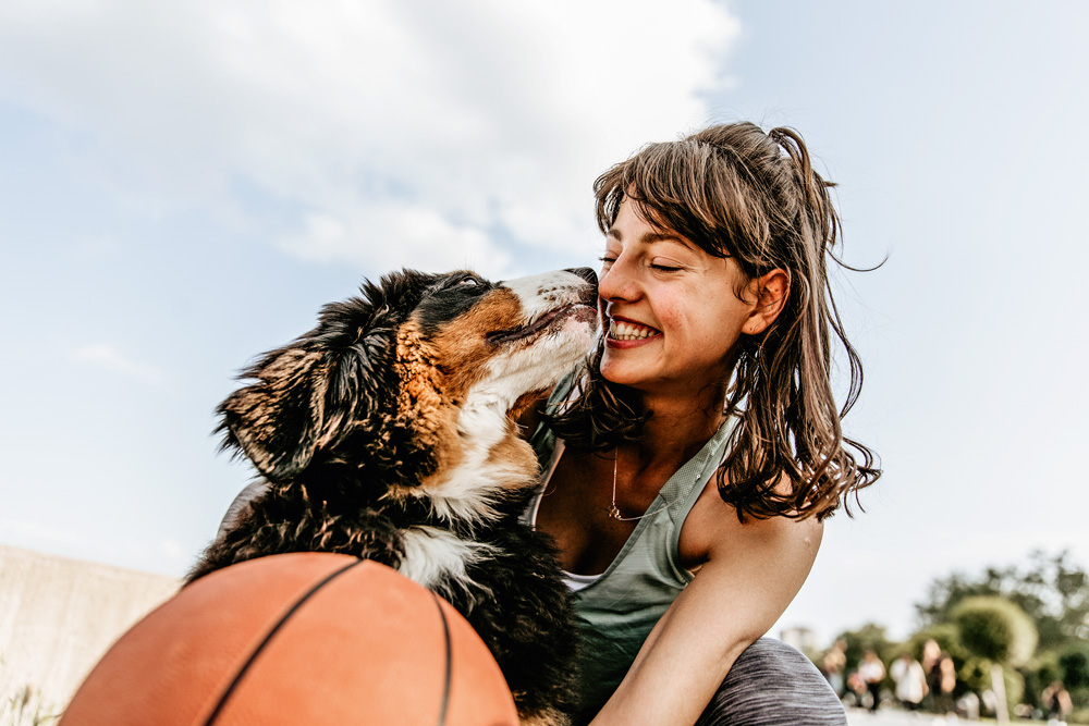 foto tirada de cima para baixo de mulher sorrindo com o cachorro, que está lambendo seu rosto. o dia está ensolarado e tem uma bola de basquete no primeiro plano