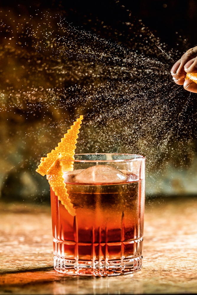 Drinque de cor alaranjada servido em um copo de fundo redondo e decorado com uma parte da casca de uma laranja