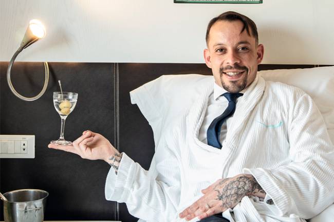 O bartender Gabriel Santana sentando sobre uma cama segurando nos dedos um martini.
