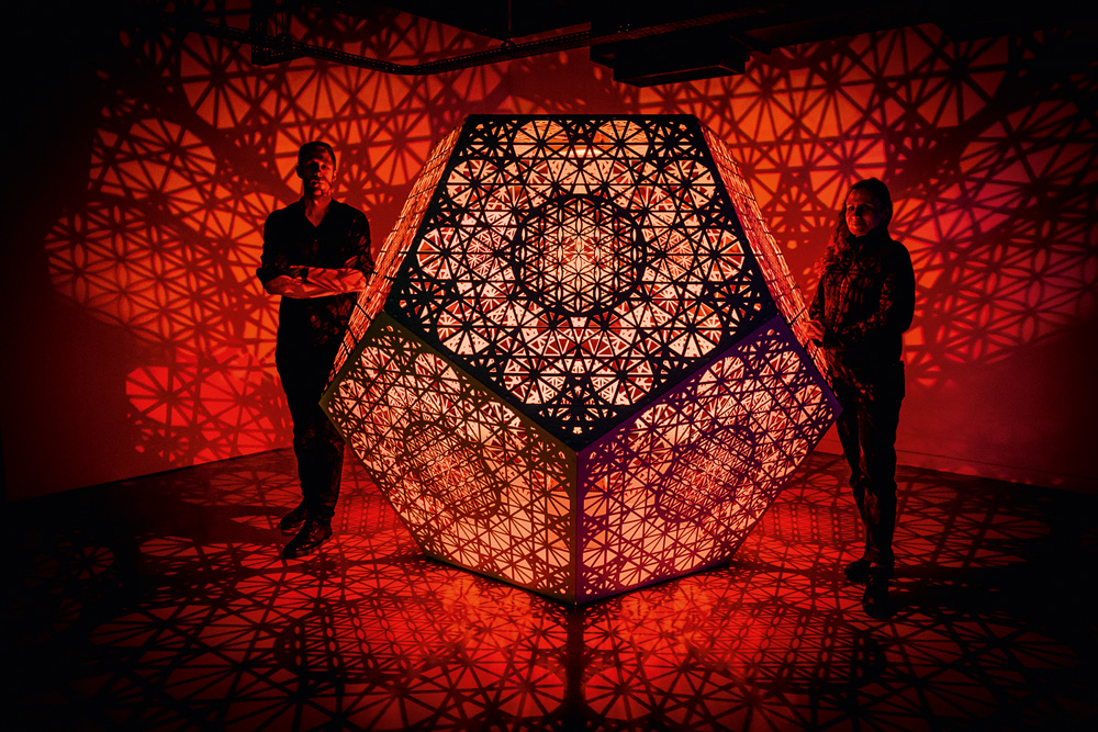 Imagem mostra octógono vazado com uma luz vermelha dentro, que faz sombras geométricas nas paredes. O quarto está escuro e duas pessoas, um homem e uma mulher, estão de cada lado da figura geométrica.