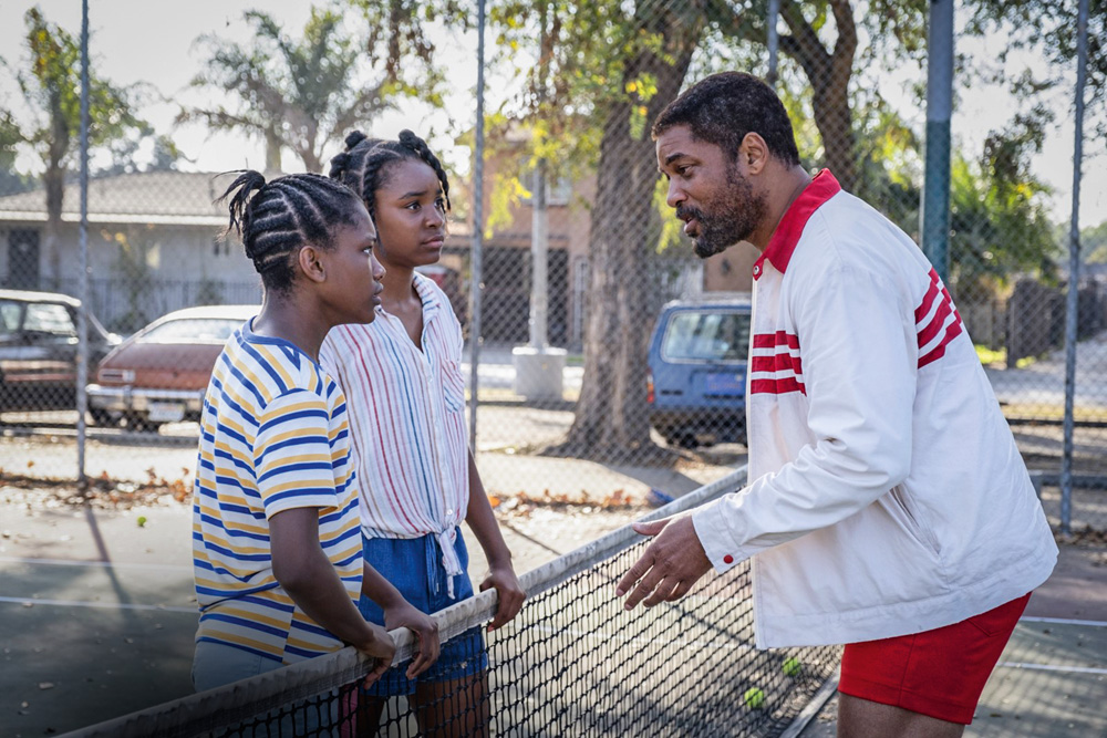Imagem mostra duas meninas de um lado da rede de tênis e um homem do outro, conversando em uma quadra.