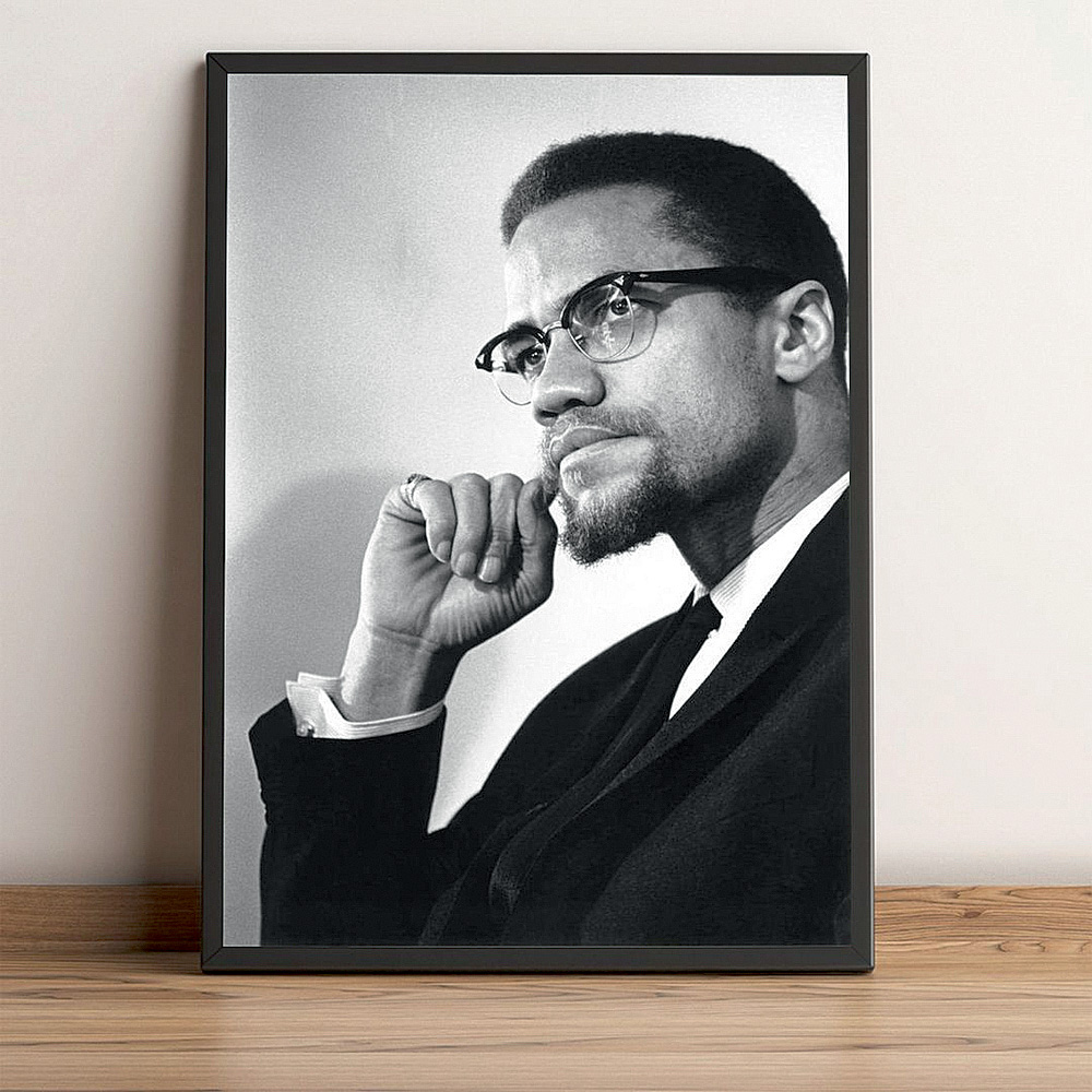 A foto mostra um quadro apoiado na parede e no chão. No quadro, uma foto em preto e branco de Malcolm X olhando para o lado e com a mão no queixo