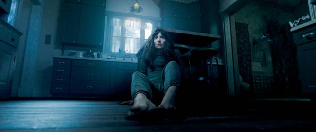 A imagem mostra uma mulher sentada no chão, com as mãos apoiadas para trás para se apoiar. Ele está com a expressão assustada em um ambiente escuro.