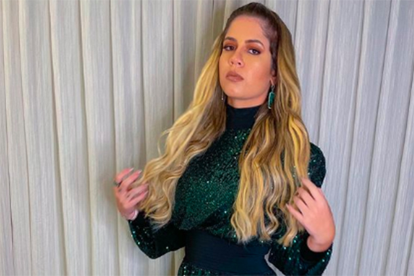 Imagem mostra Marília Mendonça em pé, fazendo pose diante de câmera, de maquiagem e vestido brilhante