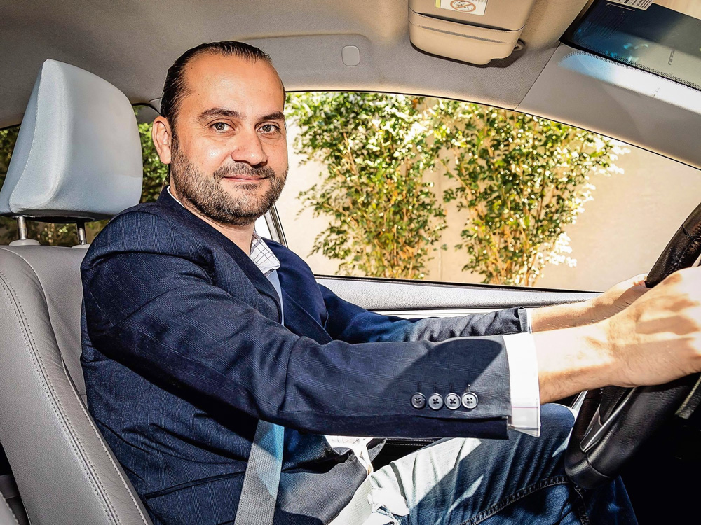 A imagem mostra Maron sentado no banco de motorista, com cinto de segurança, e mãos no volante. Ele olha para o lado, em direção a câmera, no lado do passageiro, sorrindo.