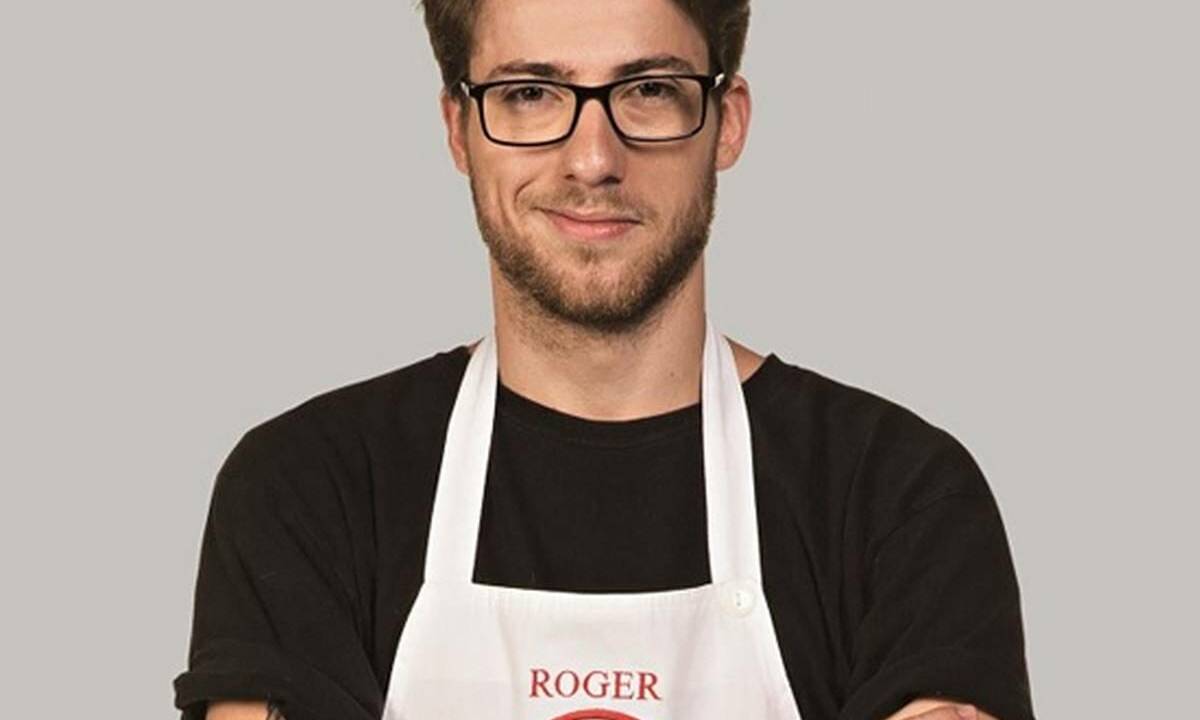 Imagem mostra Roger sorrindo, de óculos, com avental do MasterChef com o seu primeiro nome bordado