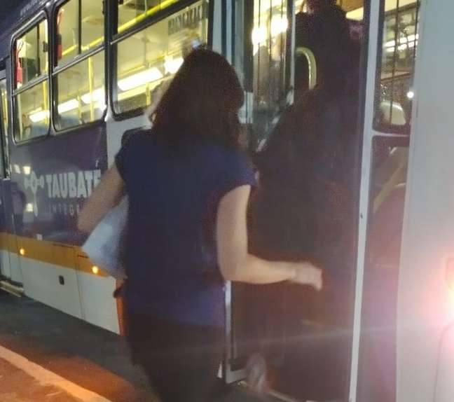 Foto mostra uma mulher de camisa azul escura e calça preta de costas subindo em um ônibus