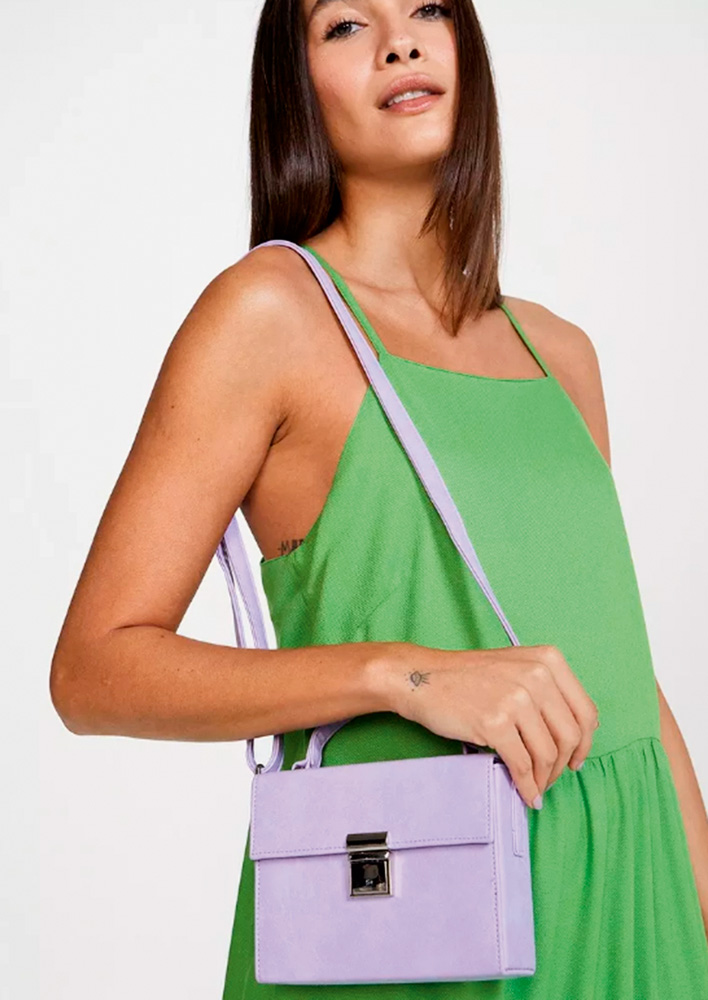 Uma mulher de vestido verde posa olhando para a foto enquanto segura uma bolsa, pendurada no ombro, lilás e quadrada