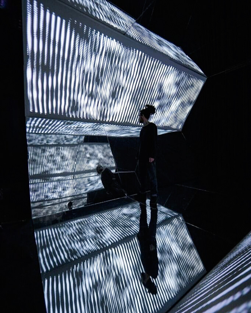 foto de VJ Vigas dentro de sua obra aparato 10, que tem múltiplas estruturas geométricas luminosas. ele está na penumbra