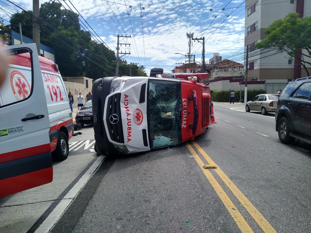Uma ambulância tombada na rua, com o capô danificado e o vidro quebrado após colisão com carro na Zona Leste de São Paulo.