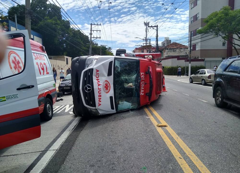 Uma ambulância tombada na rua, com o capô danificado e o vidro quebrado após colisão com carro na Zona Leste de São Paulo.
