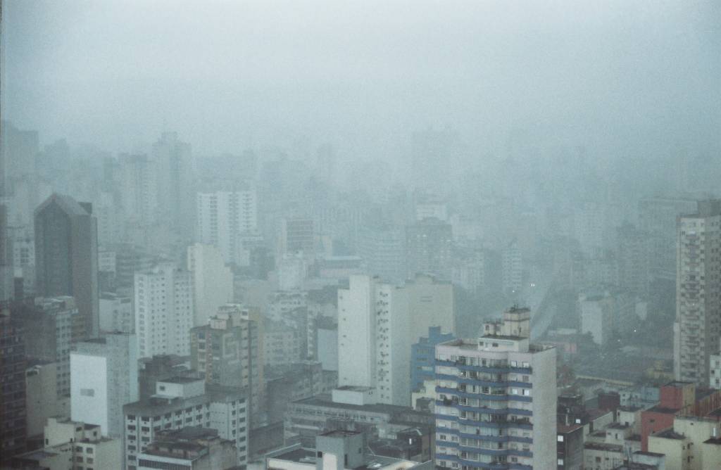 Imagem mostra horizonte de prédios sob céu nublado. Uma chuva fina ofusca a visão dos prédios.