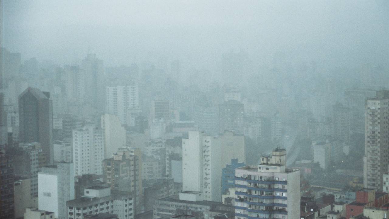 Imagem mostra horizonte de prédios sob céu nublado. Uma chuva fina ofusca a visão dos prédios.