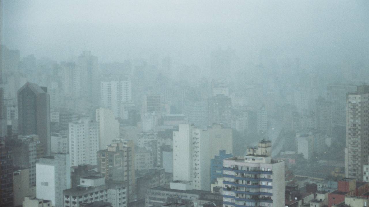 Imagem mostra horizonte de prédios sob céu nublado e chuva fraca.