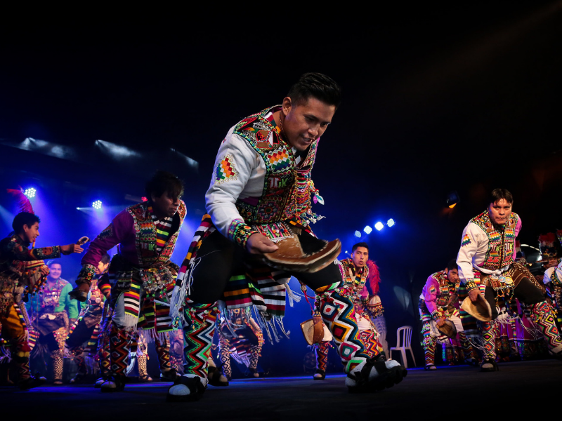 Foto de dançarinos no palco. Eles usam roupas típicas e coloridas.