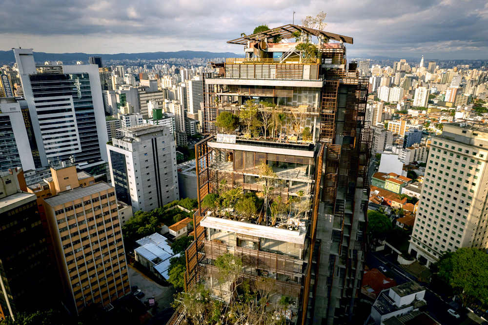 Foto aérea mostra um edifício que tem seus andares todos arborizados e com muitas plantas. Dá para ver outras dezenas de prédios ao fundo, além do céu