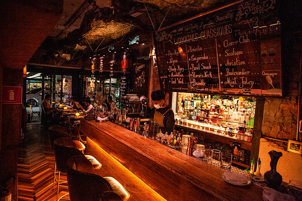 Ambiente de bar com balc&atilde;o de madeira e nomes dos drinques anotados em um vidro perto do teto