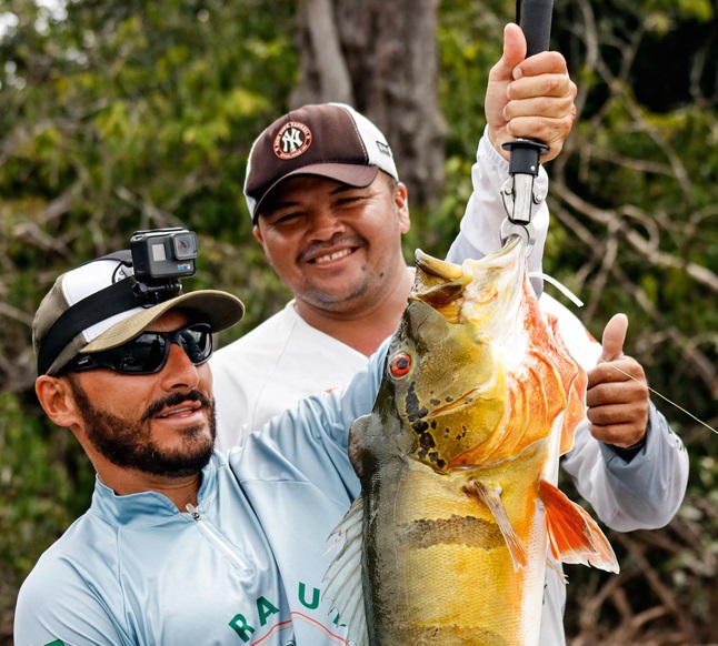 Um homem segura um grande peixe em suas mãos; Outro atrás sorri para a câmera