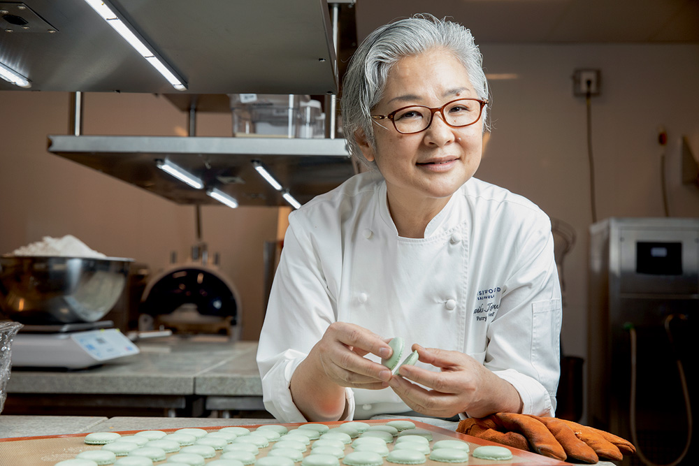 A confeiteira asiática posa apoiada em um balcão de uma cozinha de restaurante