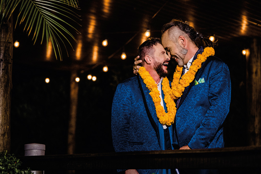 Anderson Zei (à esquerda) e Fábio Alex se abraçam com as testas encostadas. Ambos vestem ternos azuis e colares de flores laranja.