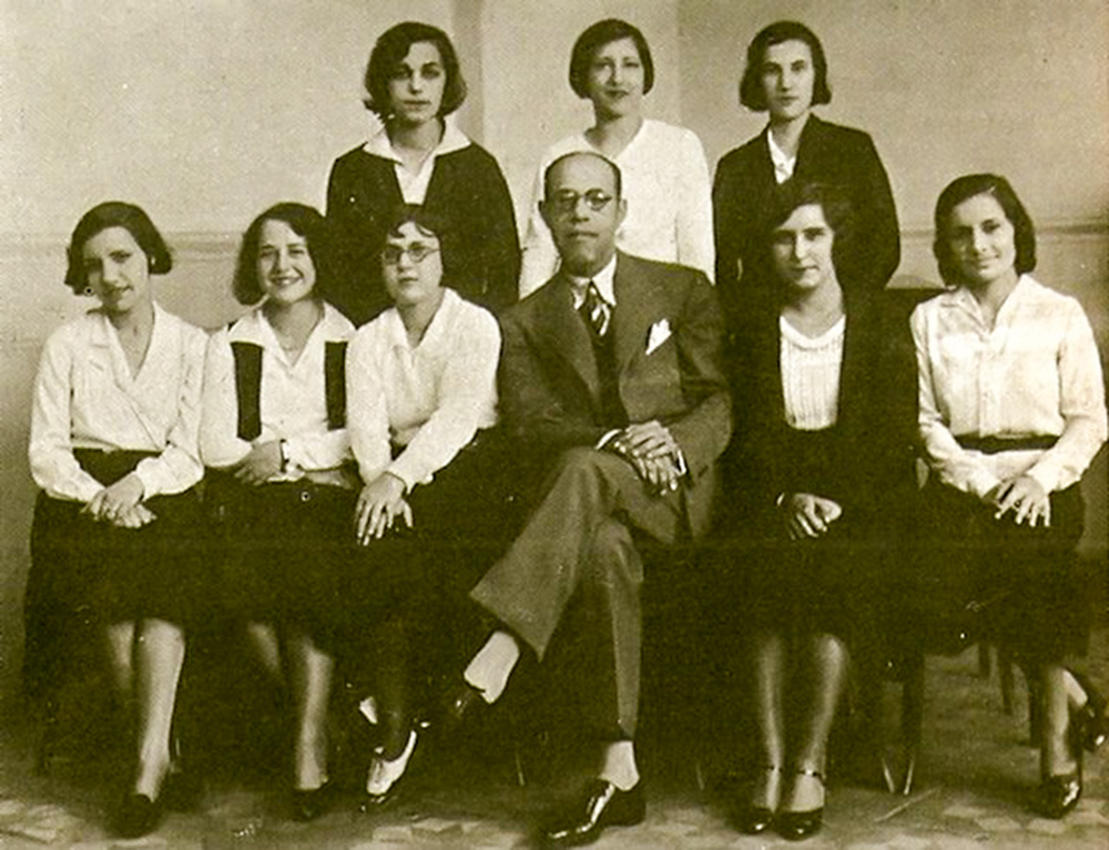 Imagem em branco e preto mostra 8 mulheres sentadas e em pé em volta de um homem, ao centro, de terno e sentado de pernas cruzadas.