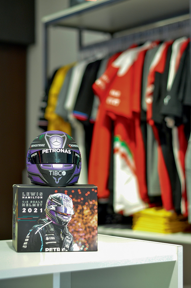 DSC_0543.jpg Foto mostra um capacete de corrida em cima de uma caixa. Ao fundo, é possível ver várias camisas de corrida