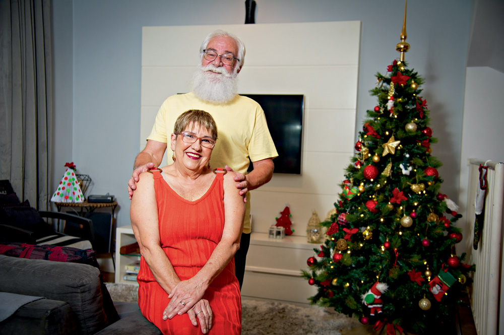 Isabel e Waldemir aparecem descaracterizados. Ela usa uma macacão vermelho e ele, uma blusa amarela. Ela está sentada e ele apoia as mãos em seus ombros por trás. Ao fundo, na sala da casa, há uma árvore de natal decorada.